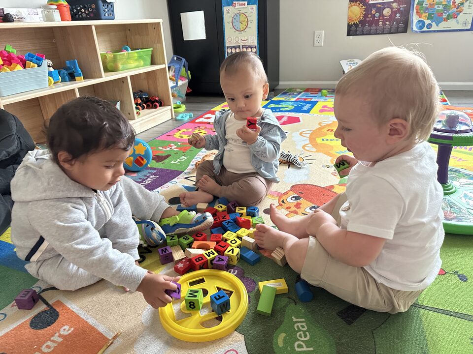 Tres bebés sentados juntos sobre una alfombra colorida del abecedario, jugando con bloques de letras de madera, con estantes de juguetes y carteles educativos detrás