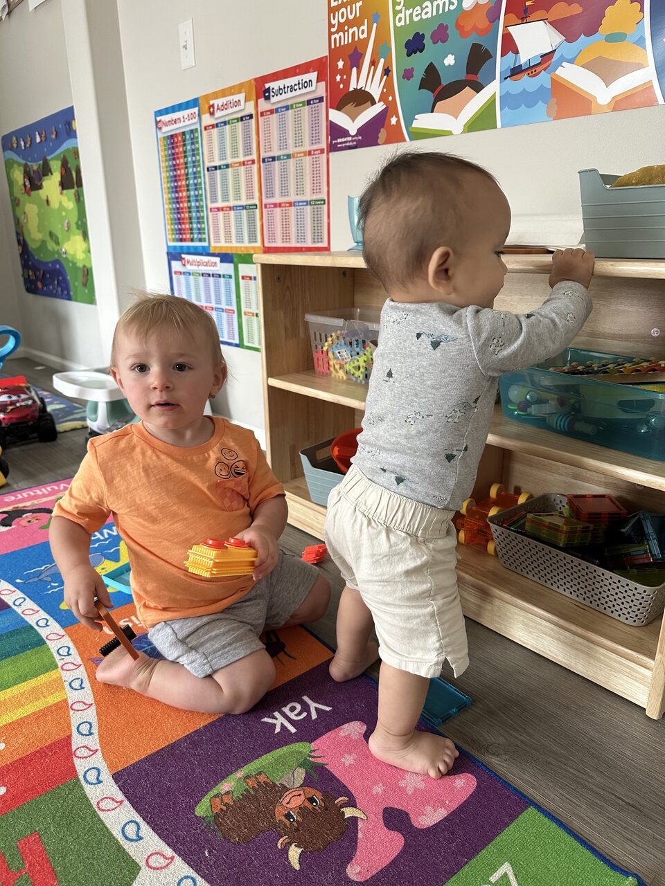 Dos niños pequeños frente a un estante bajo de madera con canastas de fichas magnéticas y juguetes, sobre una alfombra de colores y bajo carteles de letras y números